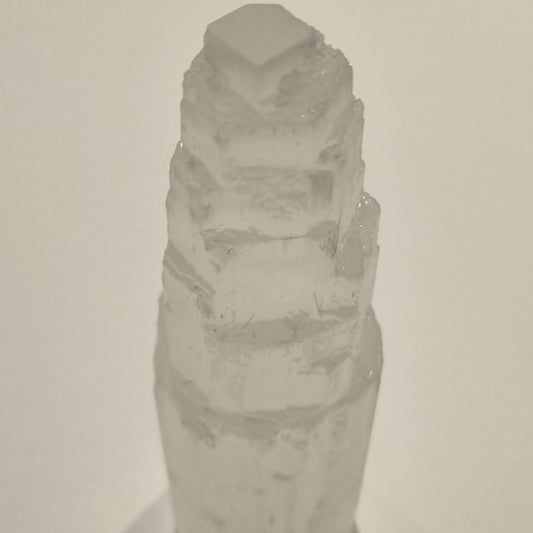 A pale translucent selenite crystal tower or geode specimen with a faceted top, photographed against a plain light background.