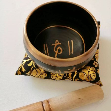Black singing bowl with gold accents on a decorative pillow, accompanied by a wooden mallet.
