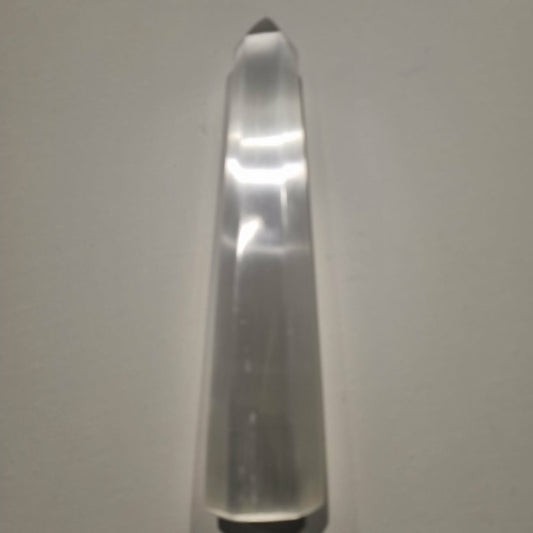 A selenite tapered crystal point, standing vertically on a plain light gray surface.
