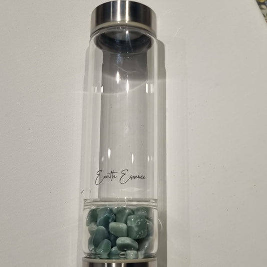 A clear cylindrical glass bottle with a silver metal cap and base, containing small green aventurine stones at the bottom. The bottle has 'Earth Essence' printed on it in black script and is photographed on a plain white surface.