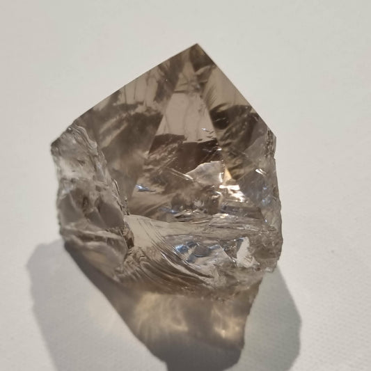 A raw, unpolished smokey quartz crystal point with natural faceted edges and a rough, textured base, photographed on a neutral light gray surface with a soft shadow.