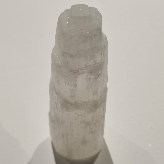 A pale translucent selenite crystal tower with a rough, layered texture, photographed against a plain light background with a soft shadow below.