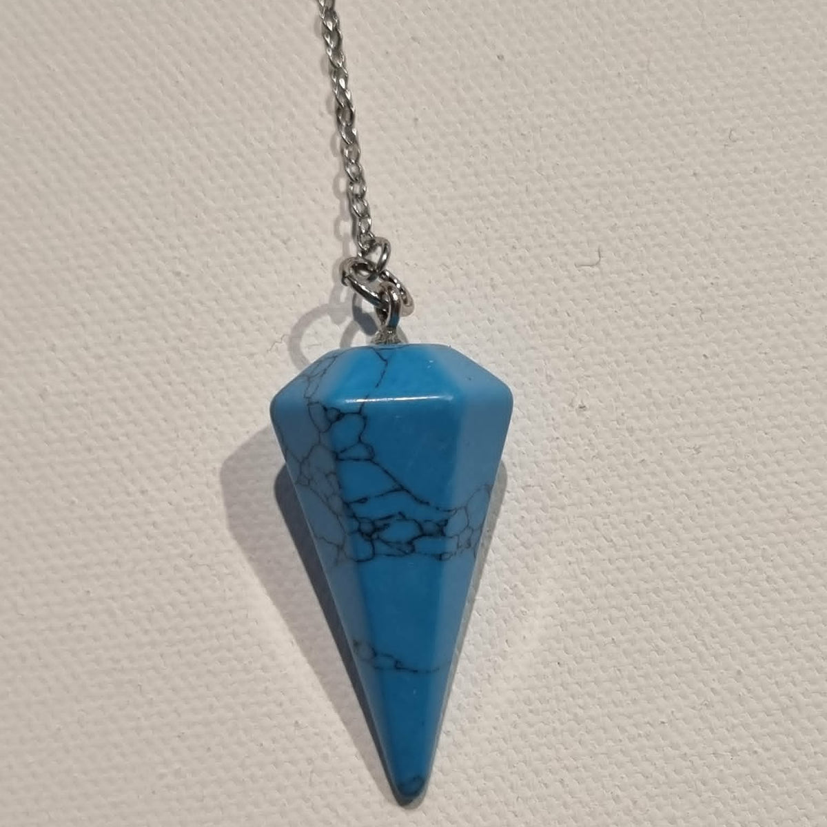 A blue howlite pendulum with natural matrix veining, suspended from a silver-toned chain against a textured off-white background.
